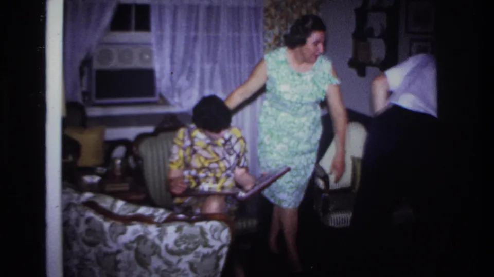 1967 domestic frame showing a woman in a green floral dress beside a seated woman in a patterned dress in a Rockaway Beach living room.