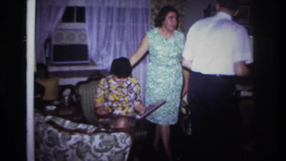 Rockaway Beach 1967 home-movie frame with adults moving and sitting in a private living room with curtains, an air conditioner, and floral furniture.