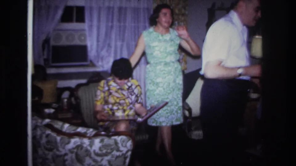 In a 1967 Rockaway Beach living room, a seated woman studies a photo album as another woman stands beside her and a man is partly visible.