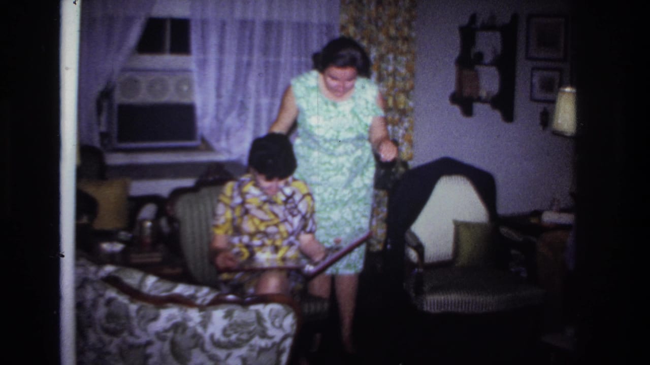 A standing woman leans toward a seated woman holding a photo album in a living room.