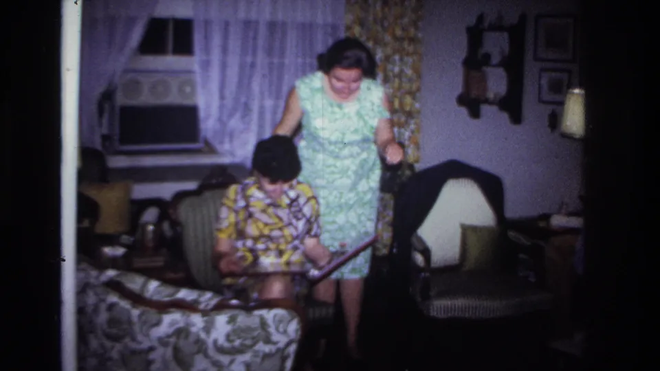 The standing woman in a green floral dress leans toward the seated woman and her photo album inside the Rockaway Beach home.