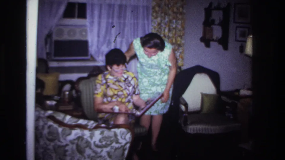 Two women examine a photo album together in a 1967 Rockaway Beach living room with period curtains, wall decor, and upholstered furniture.