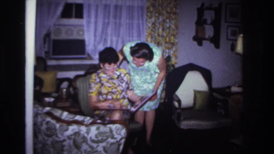 The women continue looking at the photo album in the living room, framed by a window air conditioner, curtains, shelves, and upholstered chairs.