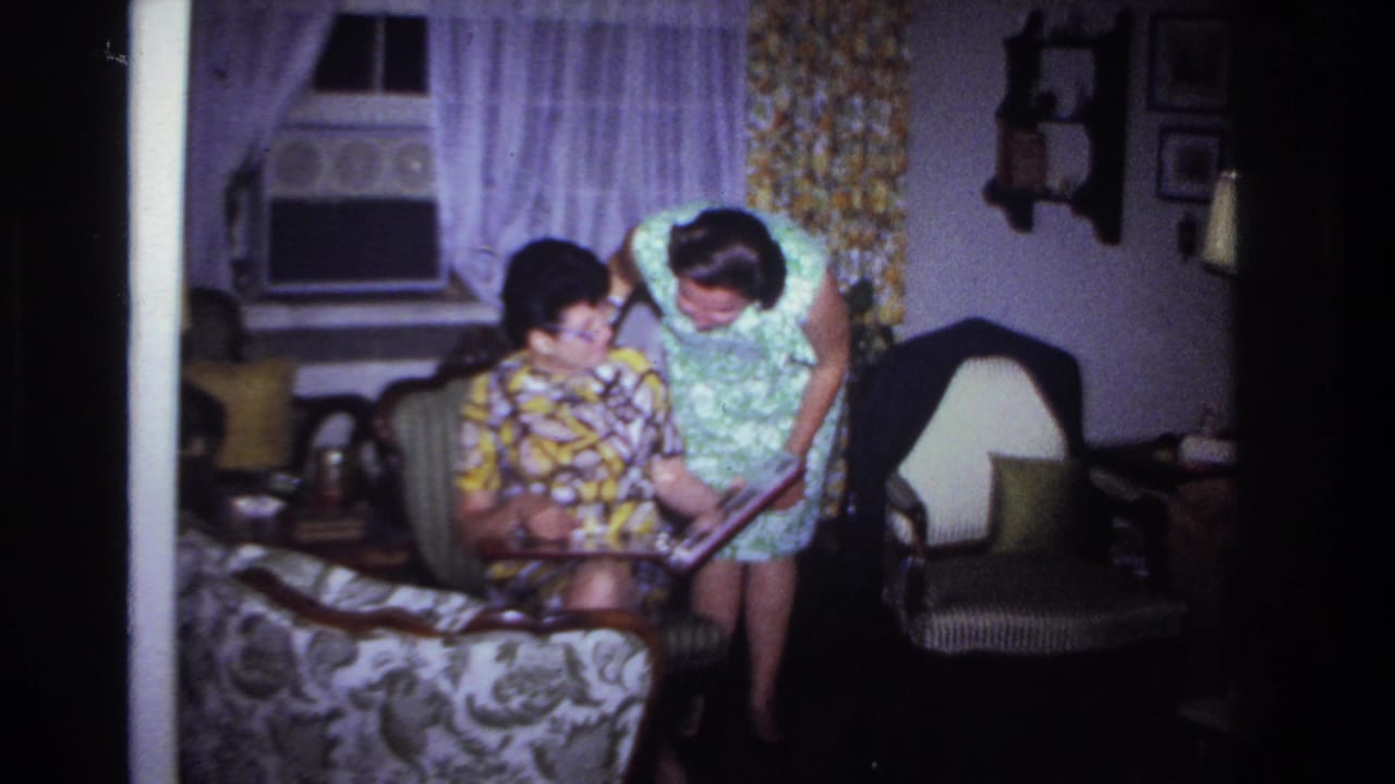 Two women in a private living room, one seated and one leaning over, look through a photo album in 1967 home-movie footage.