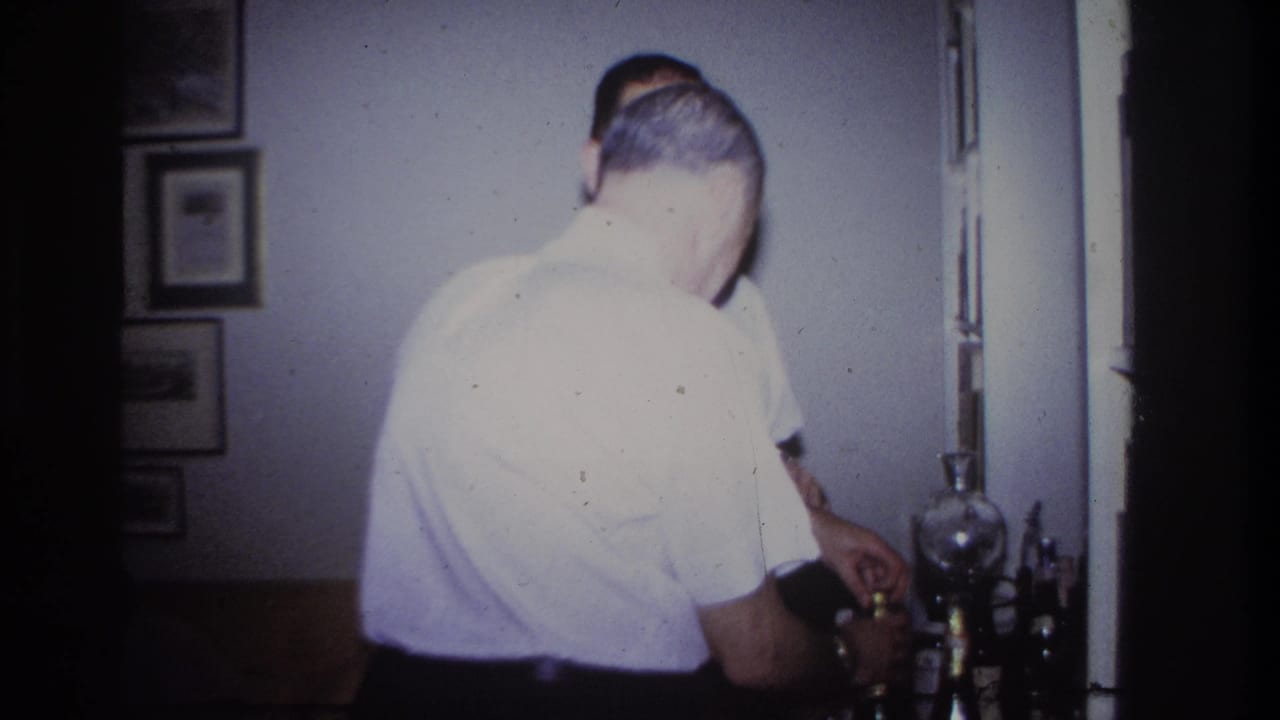 Two men in white shirts stand near a home bar with bottles and framed wall pictures.