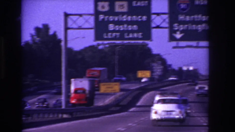A white period car travels on a divided highway beneath a Providence and Boston direction sign in 1967 road-trip footage.