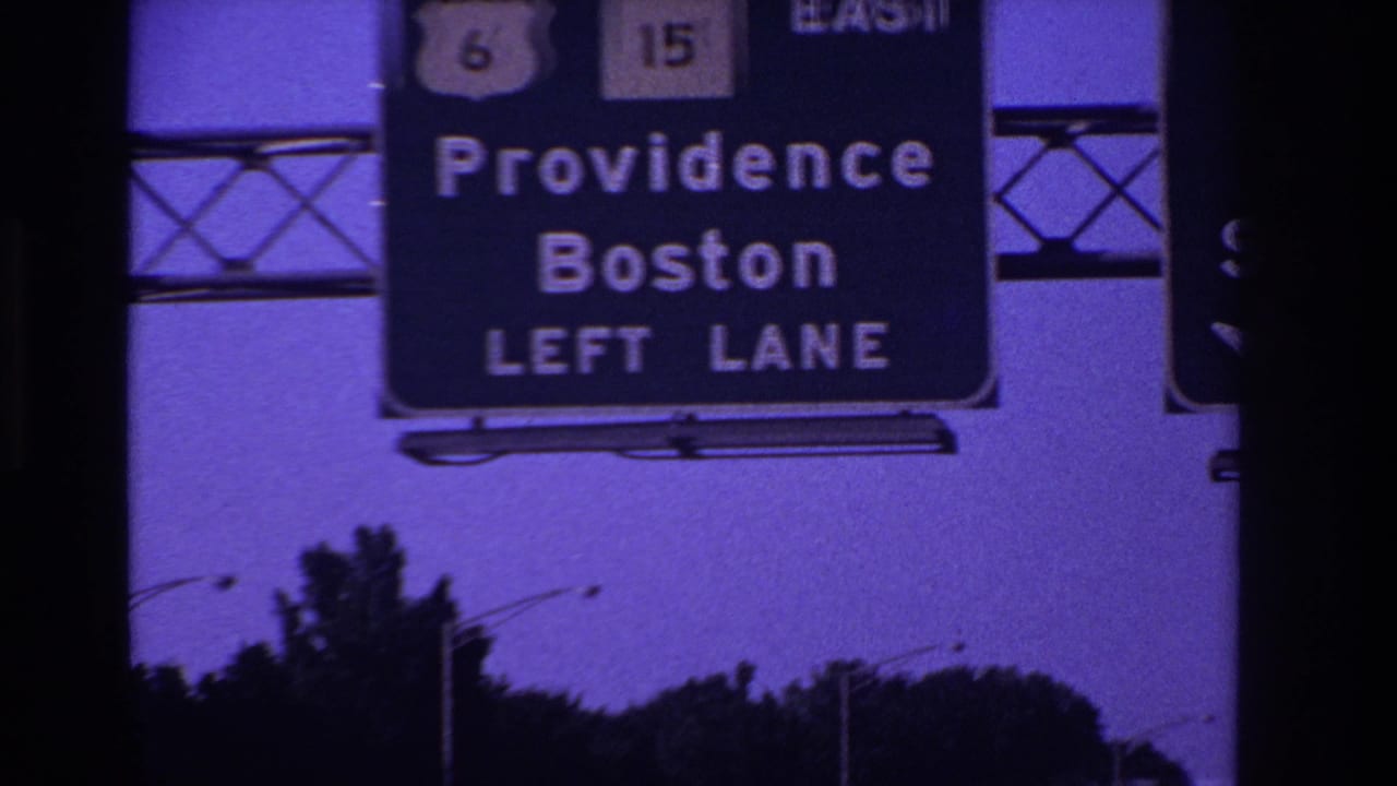 Overhead highway sign directing left-lane traffic toward Providence and Boston, seen against a purple-blue sky and roadside trees.