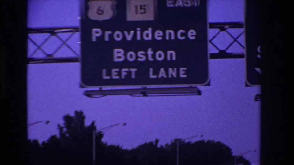 Overhead highway sign directing left-lane traffic toward Providence and Boston, seen against a purple-blue sky and roadside trees.