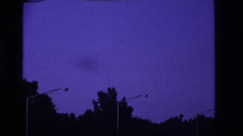 Passenger-shot 8mm frame of purple-blue sky, dark tree silhouettes, and roadside lamp posts after the car passes the sign.
