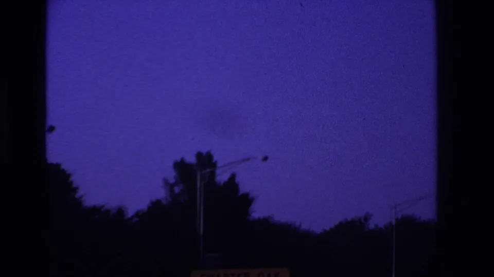 Late-clip 8mm highway view with silhouetted trees, roadside lights, purple-blue sky, and a cropped sign edge near the bottom.