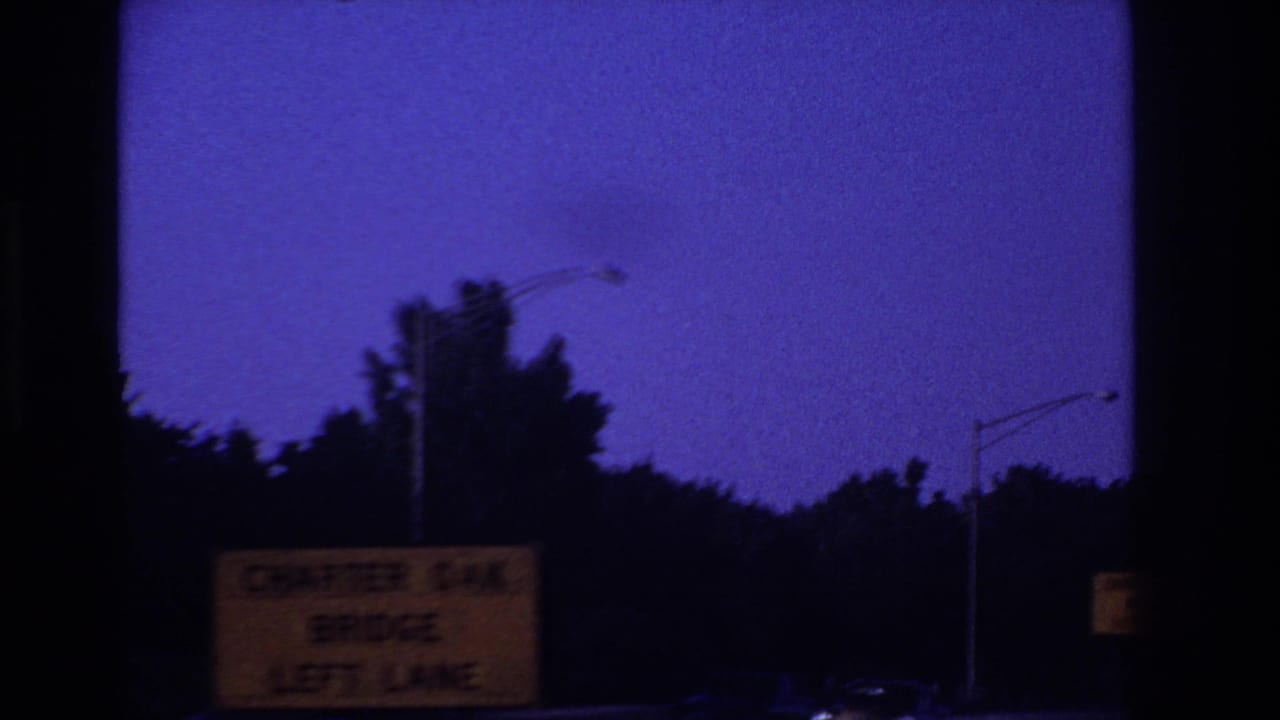 1967 8mm highway POV with a purple night sky, roadside trees, streetlights, and a partially readable Charter Oak Bridge left-lane sign.