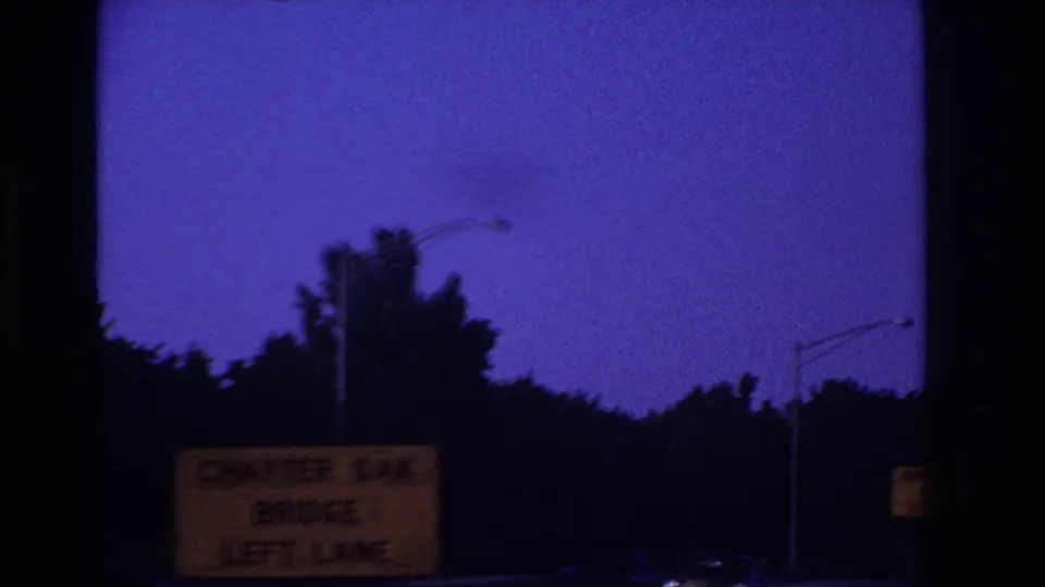 1967 8mm highway POV with a purple night sky, roadside trees, streetlights, and a partially readable Charter Oak Bridge left-lane sign.