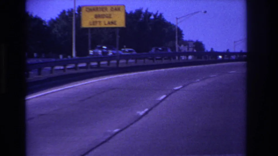 1960s 8mm driving view on a curved highway lane with bridge signage, guardrail, streetlights, and several cars in motion.