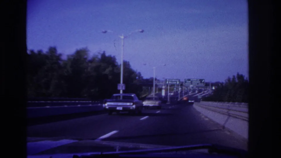 1967 home-movie driving footage shows period cars on a divided highway approaching distant green route signs.