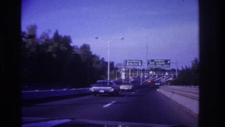Forward 8mm view of a car ahead on a northeastern U.S. highway with overhead signs and roadside trees in the distance.