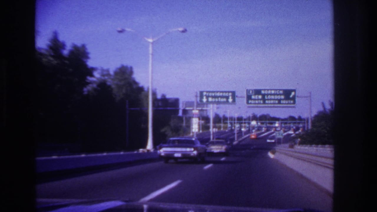 1967 8mm home-movie view from inside a moving car on a busy highway with classic cars and green destination signs ahead.