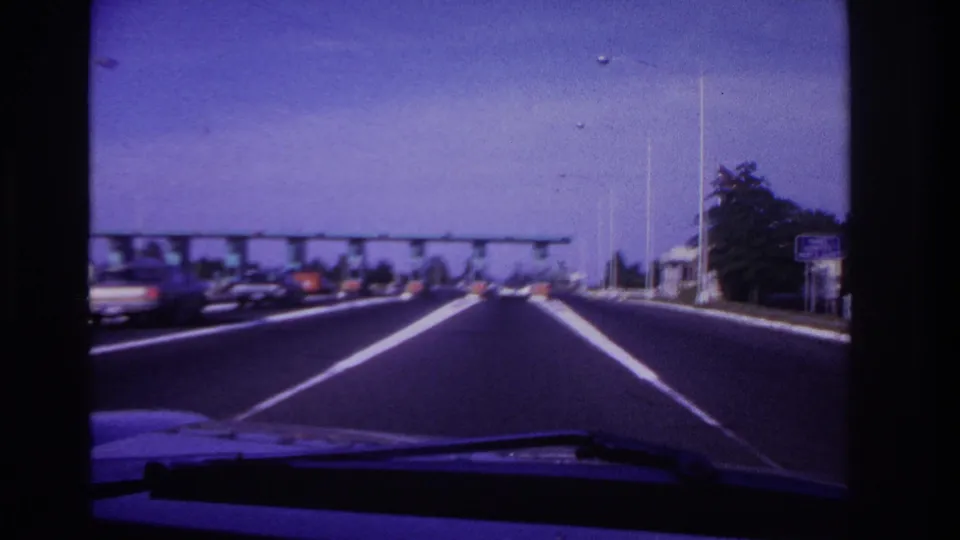 Handheld 8mm view from a car approaching a tollbooth-like highway structure with traffic, lane stripes, and roadside signs visible.