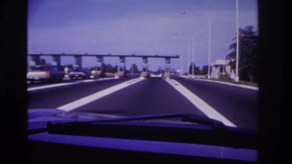 1967 8mm highway driving frame with a low dashboard edge, bright lane markings, classic cars, and an overhead gantry ahead.
