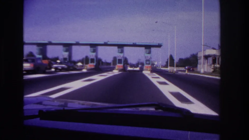 Windshield view in Rockaway Beach, 1967, nearing toll booths with period automobiles and bright lane dividers.