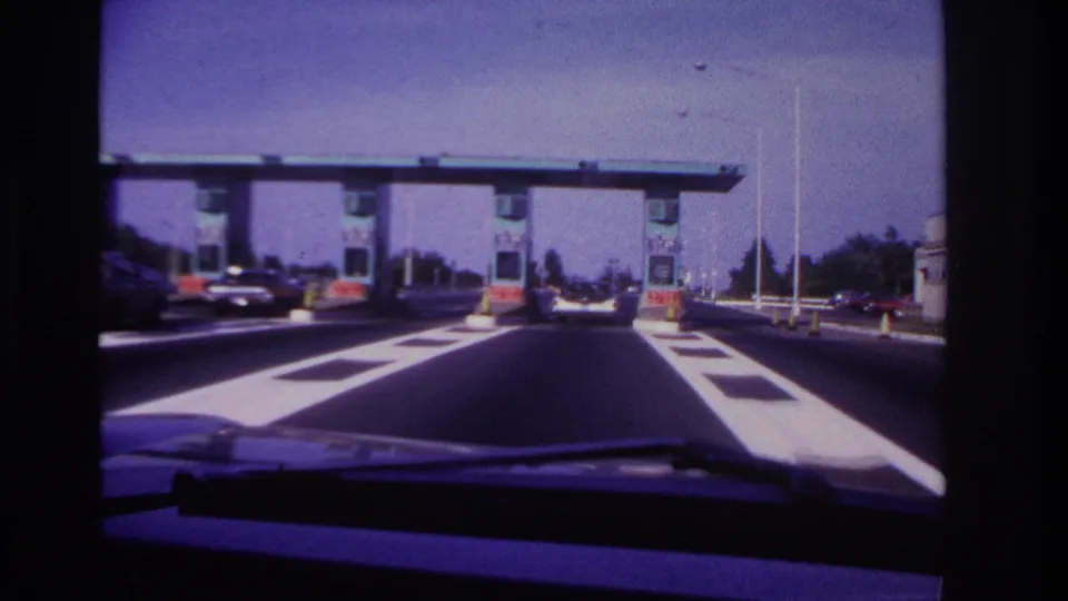 Inside-car view of a 1967 Rockaway Beach toll plaza approach, with booths, cones, and cars ahead.