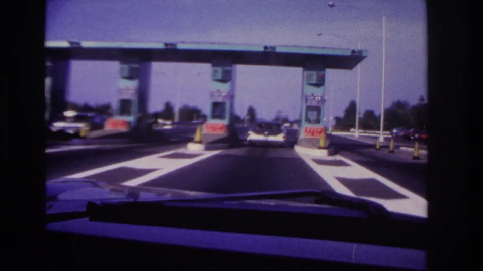 Close 1967 windshield view of a Rockaway Beach toll plaza, with period cars beside the booths and lane markings ahead.