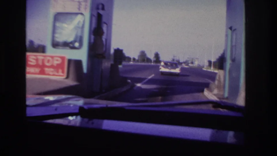 Close driver's-side view of a teal toll booth, red stop-pay-toll sign, and roadway beyond in the supplied 1967 Rockaway Beach footage.
