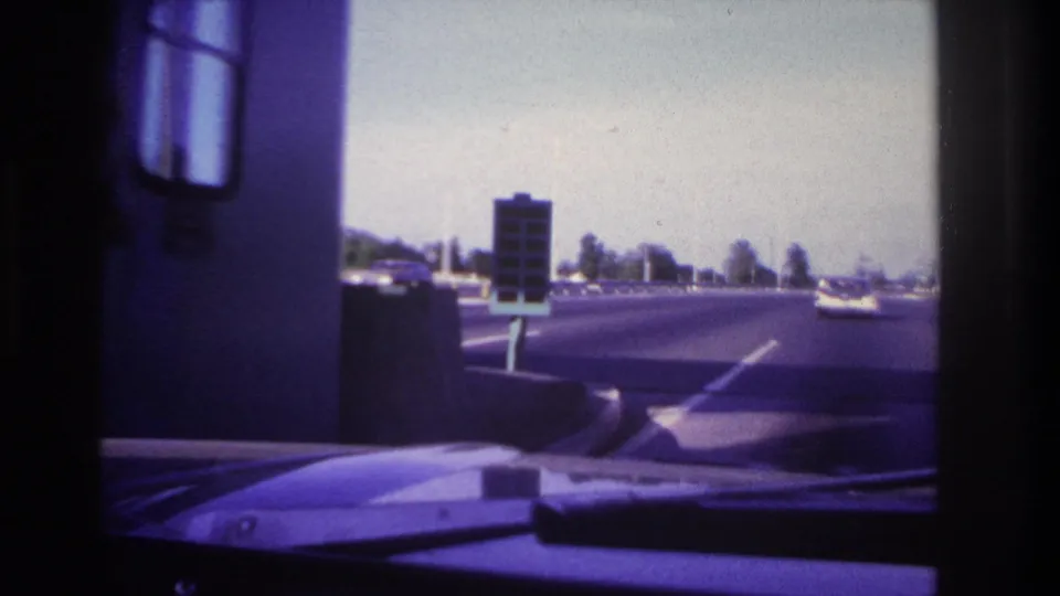 After passing the toll booth, the 1967 driver's view opens onto the roadway with lane markings, a car ahead, and dashboard visible.