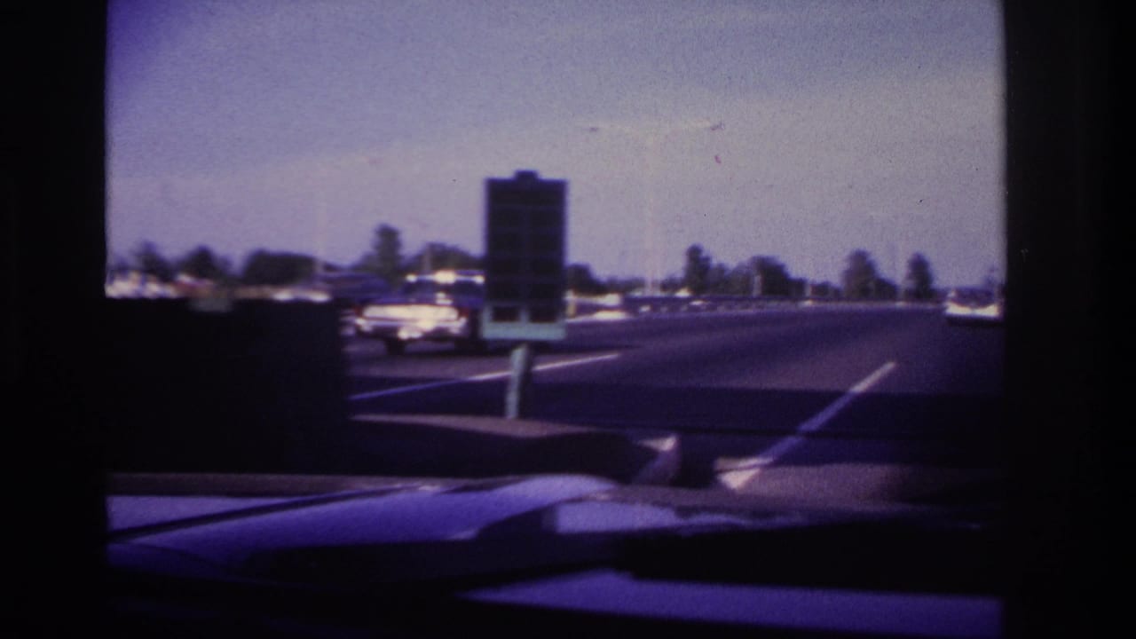 View through a car windshield of a multi-lane road with period cars and a dark roadside sign.