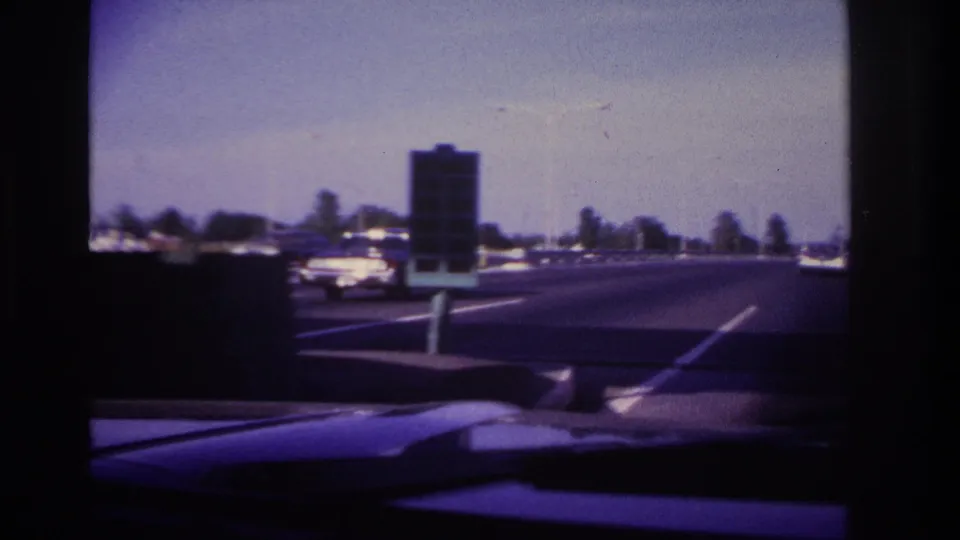 1967 Rockaway Beach road view filmed from inside a moving car, with cars ahead, lane markings, dashboard, and windshield wiper visible.