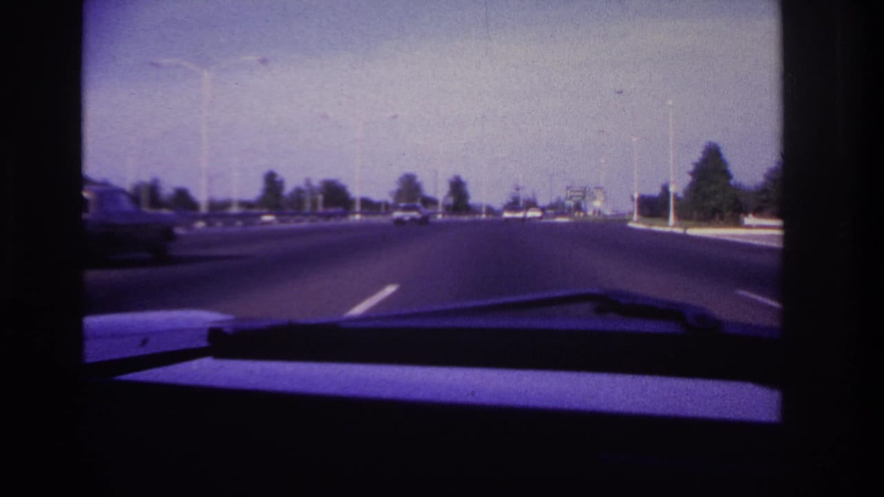 Forward-looking 1967 Rockaway Beach driving shot with a wide roadway, distant cars, tall streetlights, and the windshield wiper in foreground.