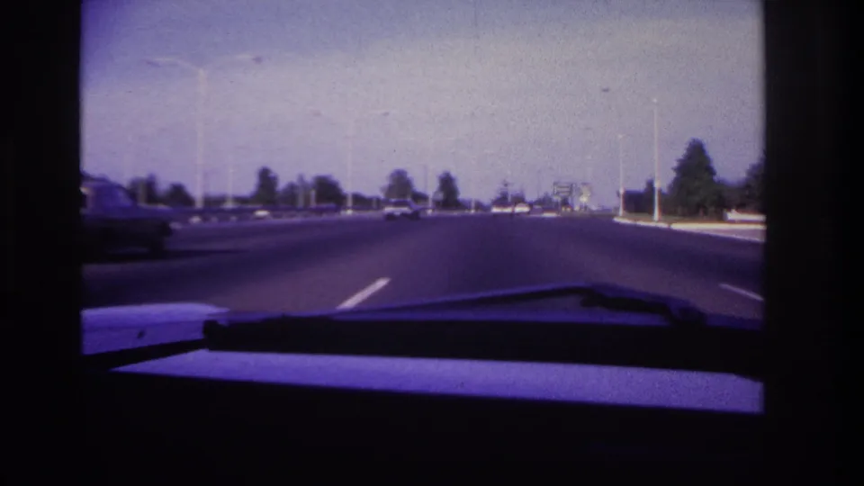 Forward-looking 1967 Rockaway Beach driving shot with a wide roadway, distant cars, tall streetlights, and the windshield wiper in foreground.