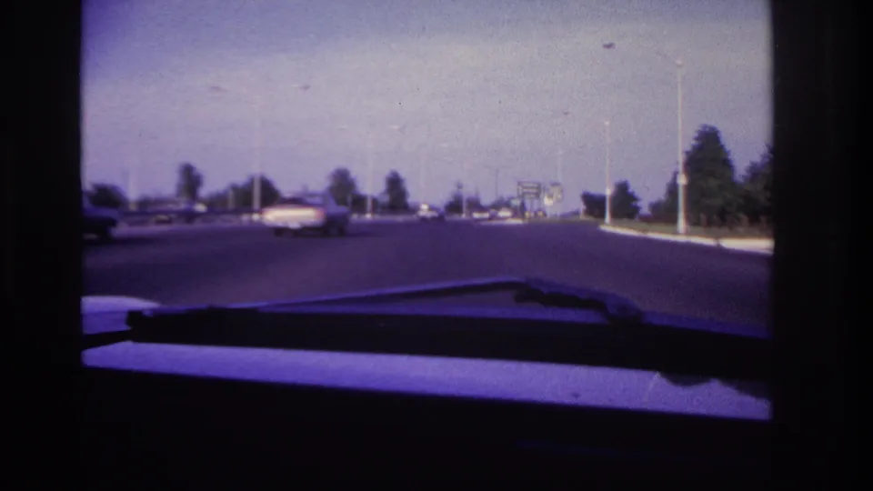 Late-1960s Rockaway Beach driving view with cars ahead on a broad road, roadside trees, streetlights, dashboard, and wiper visible.