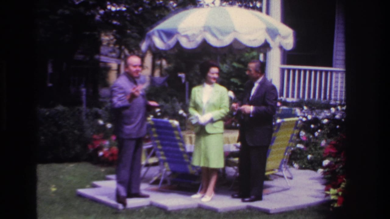Three adults in dress clothes stand in a Rockaway Beach backyard under a striped umbrella, with chairs and flowers nearby.