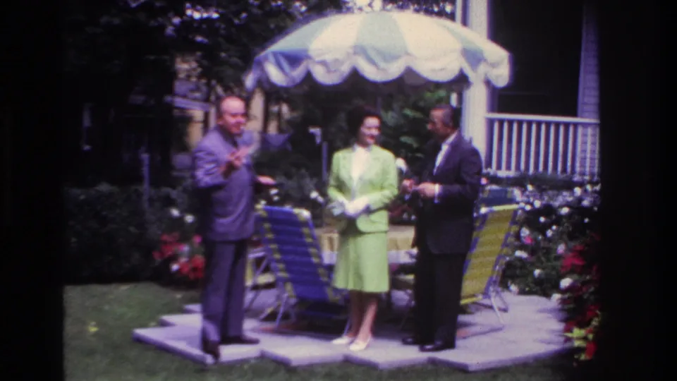Three adults in dress clothes stand in a Rockaway Beach backyard under a striped umbrella, with chairs and flowers nearby.