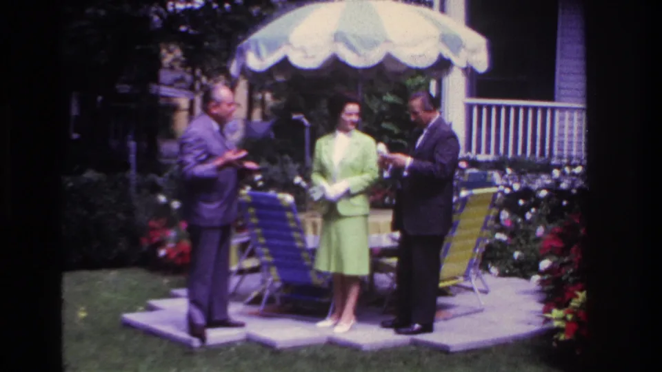 In a 1967 Rockaway Beach backyard, a woman in a green suit stands between two men near lawn chairs and flower beds.