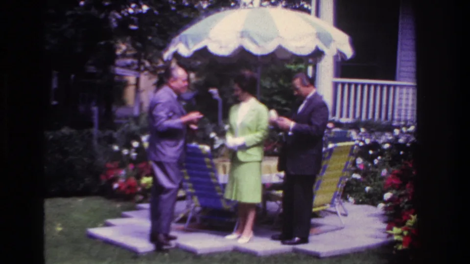 The backyard group continues standing under the striped umbrella, with porch railing, flowers and folding chairs visible.