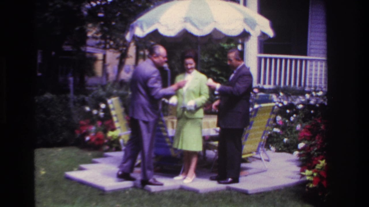 In 1967 Rockaway Beach home-movie footage, two suited men and a woman in a lime-green suit stand by lawn chairs under a patio umbrella.