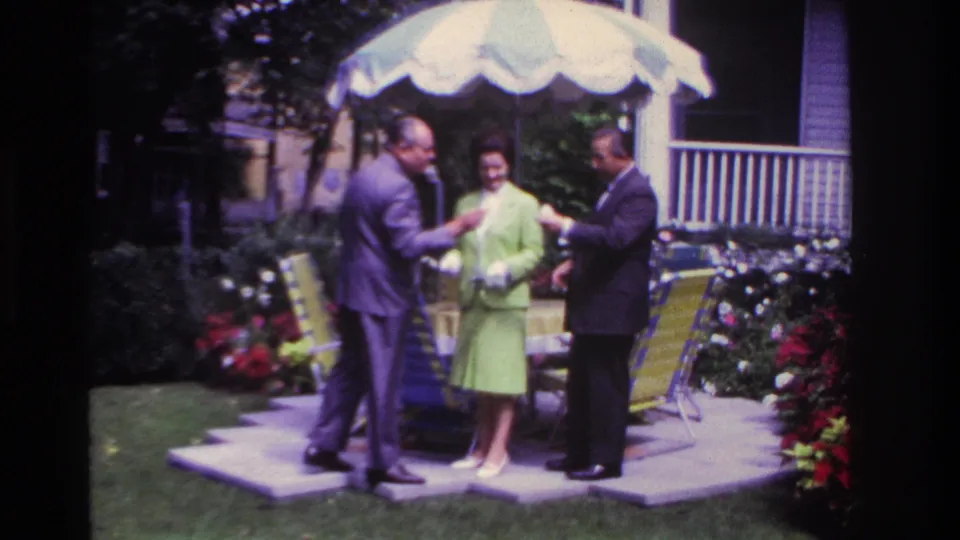 In 1967 Rockaway Beach home-movie footage, two suited men and a woman in a lime-green suit stand by lawn chairs under a patio umbrella.