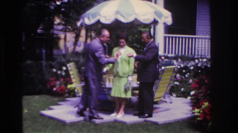 The Rockaway Beach backyard scene shows three adults gathered closely under a striped umbrella with lawn chairs and flowers behind them.