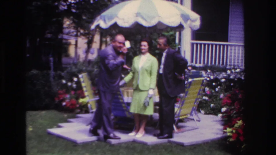 In the 1967 home-movie frame, a suited man gestures with a paper while a woman in green and another suited man watch on the patio.