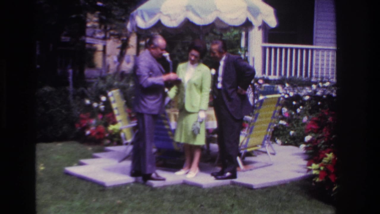 Rockaway Beach, 1967: a suited man holds out a pamphlet as a woman in green and another suited man watch on a garden patio.