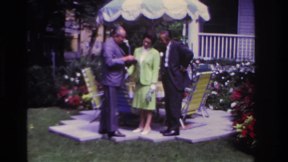 Rockaway Beach, 1967: a suited man holds out a pamphlet as a woman in green and another suited man watch on a garden patio.