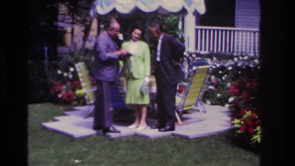 A 1967 8mm frame shows three adults in formal clothing gathered on a Rockaway Beach patio beside flowers, chairs, and an umbrella.