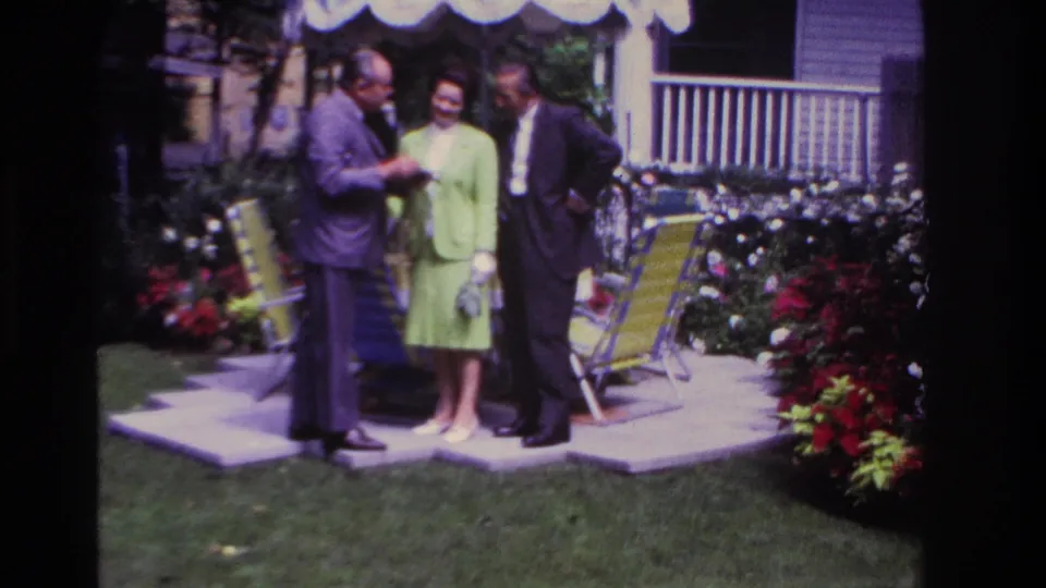 In the final 1967 Rockaway Beach frame, the woman in green faces the camera as two suited men stand near her in a backyard garden.