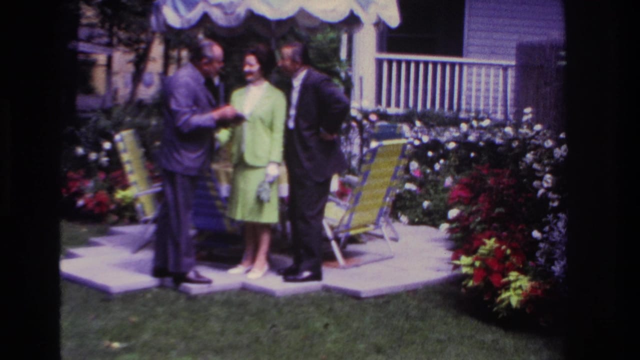 1967 Rockaway Beach home-movie frame showing two men and a woman in a lime-green suit conversing in a garden beside lawn chairs.
