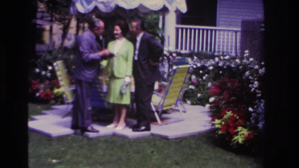 1967 Rockaway Beach home-movie frame showing two men and a woman in a lime-green suit conversing in a garden beside lawn chairs.