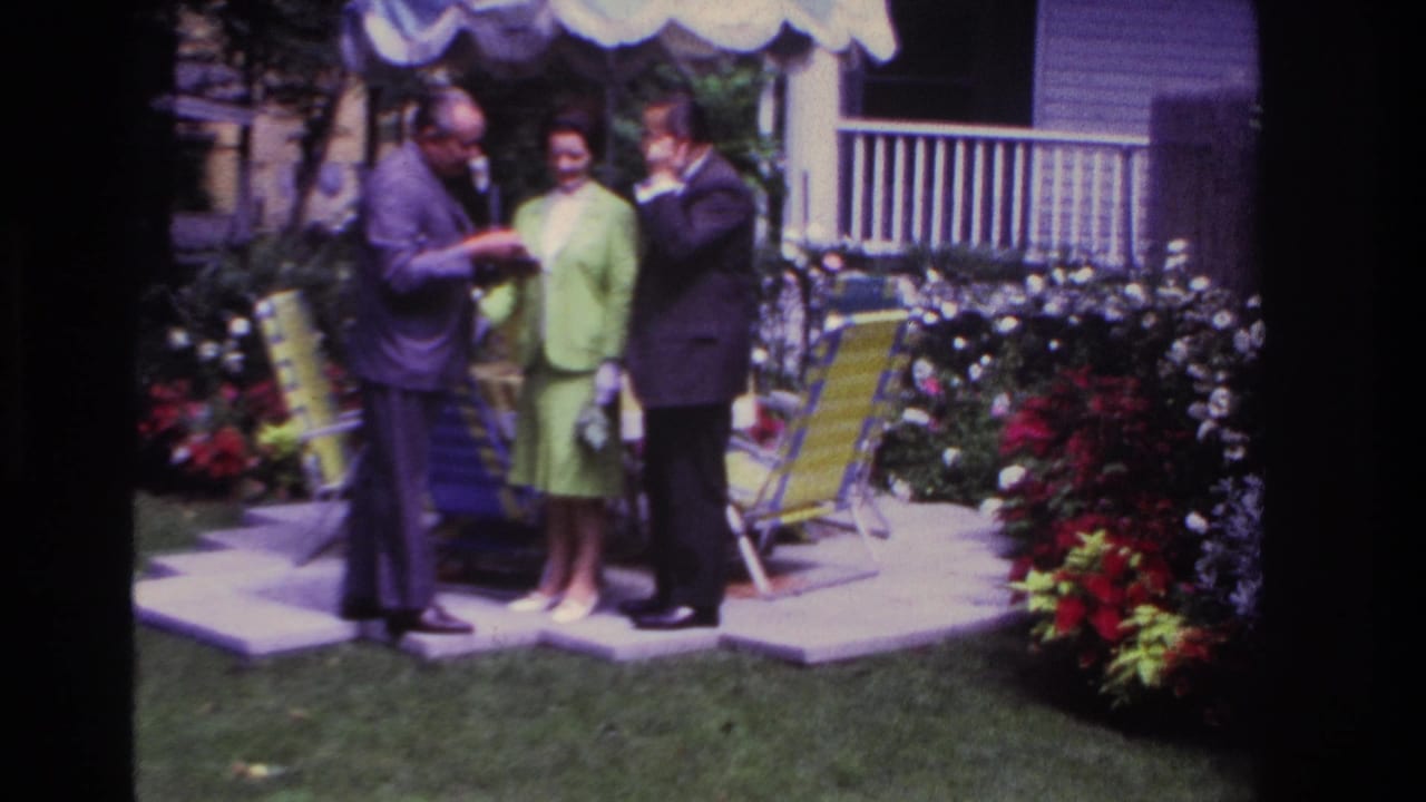 Two men and a woman stand together on a backyard patio near flowers and lawn chairs.