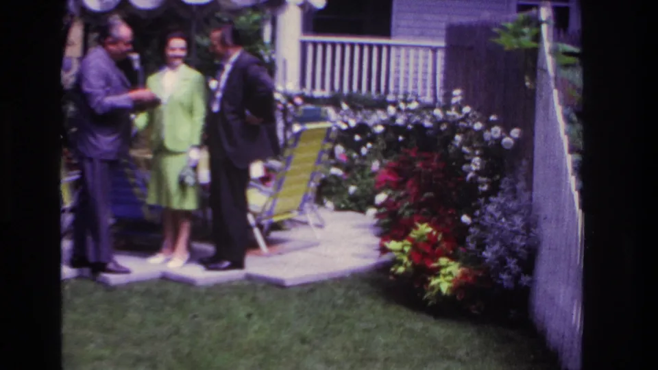 Home-movie frame from Rockaway Beach in 1967 showing adults talking in a residential garden with chairs, flowers, and a shaded porch.