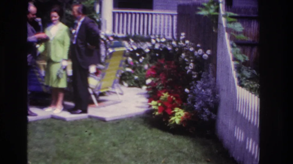 1967 Rockaway Beach 8mm frame showing an adult group in a backyard garden, with a fence, lawn chairs, porch rail, and flowers around them.