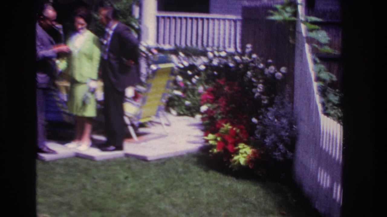 1967 Rockaway Beach home-movie frame showing three adults in dress clothes near lawn chairs, flower beds, a porch, and a striped umbrella.